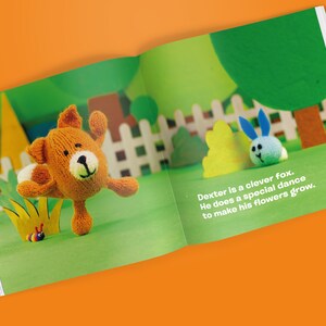 May include: A page from a children's book showing a knitted fox and a blue bunny on a green background. The fox is orange with a white belly and is dancing. Text reads: "Dexter is a clever fox. He does a special dance to make his flowers grow."