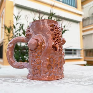 May include: A handcrafted brown ceramic mug with a distinctive, textured design. The mug has a large handle and a series of holes and raised circular details on the body. The mug is placed on a white surface.