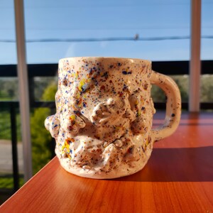 Puede incluir: Una taza de cerámica moteada con una superficie texturizada y un asa. La taza es de color blanquecino con motas coloridas de azul, amarillo y rojo. La taza está sobre una superficie de madera.