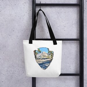 Puede incluir: Bolsa de tela blanca con asas negras que presenta un gráfico de Half Dome en el Parque Nacional de Yosemite. El gráfico tiene la forma de una punta de flecha marrón.