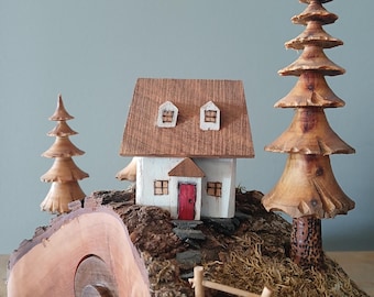 May include: A handcrafted wooden miniature village scene featuring a white house with a brown roof and a red door, flanked by carved wooden trees. The scene is set on a base of bark and moss, with a small wooden fence.