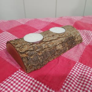 May include: A rustic, wooden candle holder with two tea light candles. The candle holder is made from a section of a log, featuring the natural bark texture. The candles are white, and the holder sits on a red and white checkered surface.