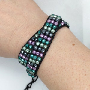 May include: A multi-strand bracelet featuring rows of small, round beads in shades of green, purple, and gray. The beads are strung on a black cord, creating a woven effect. The bracelet is tied with a black cord.