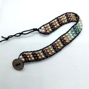 May include: Handmade beaded bracelet featuring a button closure and a woven design. The bracelet showcases a pattern of brown, cream, and teal beads, strung on a black cord. The bracelet is approximately 7 inches long.