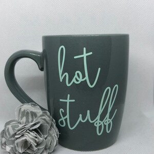 May include: A gray ceramic mug with a white handle. The mug has the words "hot stuff" written on it in a light green color.