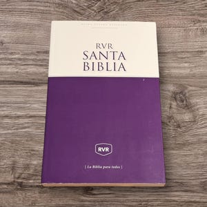 Op de afbeelding: Een hardcover boek met een witte en paarse omslag. De titel "SANTA BIBLIA" is gedrukt in grote, zwarte letters. Het boek bevat ook de tekst "RVR" en "[La Biblia para todos]" op de omslag.