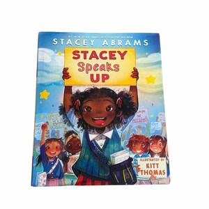 May include: A children's book cover featuring a smiling Black girl holding a sign that reads "STACEY Speaks UP." The book is by Stacey Abrams and illustrated by Kitt Thomas. The background shows a crowd of children with signs.