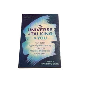 May include: A book titled "The Universe is Talking to You" with a dark blue cover and white text. The cover features a watercolor design in shades of blue and teal. The book's subtitle reads "Tap into Signs & Synchronicity to Reveal Magical Moments Every Day."
