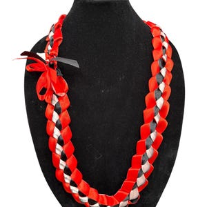 May include: A braided necklace made of red, black, and silver satin ribbons. The necklace is displayed on a black mannequin. A red bow and black ribbon accent the top of the necklace.