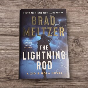 Puede incluir: Un libro de tapa dura titulado "The Lightning Rod" de Brad Meltzer. La portada presenta una silueta contra un cielo tormentoso con rayos. El título y el nombre del autor están en letras doradas.