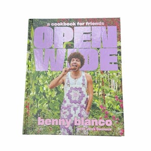 May include: A cookbook titled "OPEN WIDE" by Benny Blanco with Jess Damuck. The cover features a person in a white tank top and purple overalls, holding food. The background is a garden with green foliage and red flowers.
