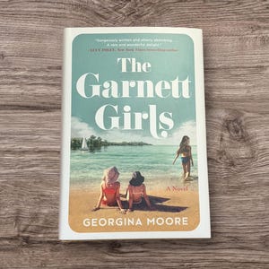 Pode incluir: Um livro de bolso intitulado "The Garnett Girls" de Georgina Moore. A capa apresenta uma cena de praia com duas figuras em trajes de banho e uma menina na água. O livro tem uma borda branca e uma citação de Lucy Foley.
