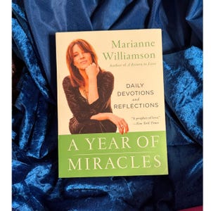 May include: A book titled "A Year of Miracles" by Marianne Williamson. The cover features a photo of the author and the text "Daily Devotions and Reflections." The book is set against a blue velvet background.