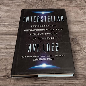 May include: A hardback book titled "Interstellar" by Avi Loeb. The cover is black with white text and a graphic of a blue and white planet. The book's subtitle reads "The Search for Extraterrestrial Life and Our Future in the Stars."