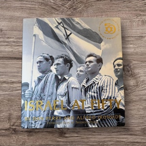 Könnte beinhalten: Ein Buch mit dem Titel "Israel at Fifty" mit einem Foto einer Gruppe von Männern und einer Flagge. Das Cover hat goldene Schrift und ein goldenes Emblem. Der Hintergrund ist eine Holzoberfläche.