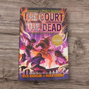 Può includere: Un libro intitolato "The Court of the Dead: A Nico Di Angelo Adventure" di Rick Riordan e Mark Oshiro. La copertina presenta un'illustrazione di personaggi e una figura cornuta in tonalità viola e arancioni. È visibile un adesivo esclusivo di Barnes & Noble.