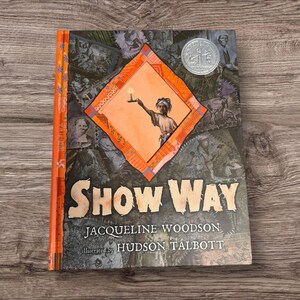 May include: A hardcover book titled "Show Way" by Jacqueline Woodson, illustrated by Hudson Talbott. The cover features a central orange diamond with a figure holding a candle, surrounded by a collage of images. A silver coin is visible near the top.