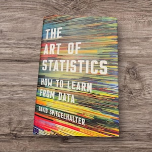 Pode incluir: Um livro de capa dura intitulado "The Art of Statistics: How to Learn from Data" de David Spiegelhalter. A capa apresenta um design abstrato colorido com linhas horizontais em tons de azul, verde, amarelo e vermelho. O título está em letras brancas grandes.