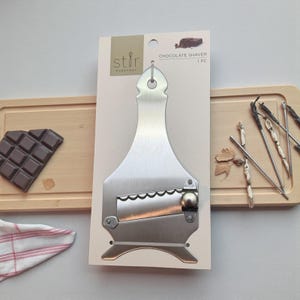 Puede incluir: Un afeitadora de chocolate de acero inoxidable con una cuchilla dentada, presentada en una tarjeta blanca con el texto "Chocolate Shaver". La tarjeta está sujeta a una tabla de cortar de madera. También hay una tableta de chocolate.