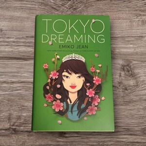 Könnte beinhalten: Ein Buch mit dem Titel "TOKYO DREAMING" von Emiko Jean. Das Cover ist grün und zeigt eine Frau mit Tiara, umgeben von rosa Kirschblüten. Das Buch ist von der New York Times Bestsellerautorin von TOKYO EVER AFTER.