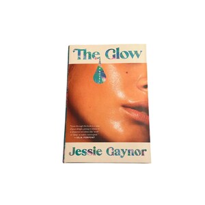 May include: A paperback book titled "The Glow" by Jessie Gaynor. The cover features a close-up of a person's face with glowing skin. The title is in teal and white, with a teal teardrop graphic. A quote is printed on the cover.