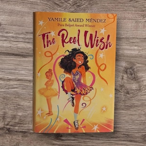 Könnte beinhalten: Ein Buch mit dem Titel "The Reel Wish" von Yamile Saied Méndez. Das Cover zeigt eine tanzende Mädchen in einem bunten Kleid, mit einer Ballerina im Hintergrund. Der Hintergrund ist gelb mit orangefarbenen Wirbeln und Sternen.