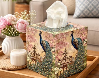 May include: A square tissue box cover featuring a printed design of two peacocks perched on a black wrought iron fence. The background displays soft pink flowering trees and white blossoms. A white tissue is pulled through the top opening.