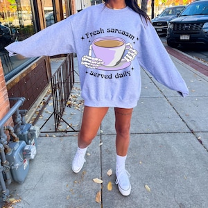 Peut inclure: Un sweat-shirt gris clair avec le texte "Fresh sarcasm served daily" et un dessin d'une tasse à café tenue par des mains de squelette. La tasse et la soucoupe sont violet clair. Le sweat-shirt a des manches longues.