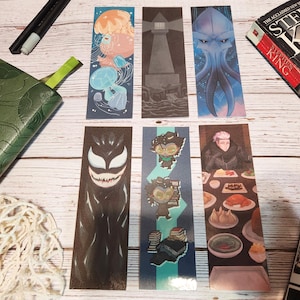 May include: A collection of six illustrated bookmarks featuring various designs. The bookmarks depict images of jellyfish, a lighthouse, an alien, a comic book character, anime characters, and a person at a table. A green leather journal and a book are also visible.