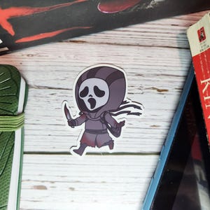 Pode incluir: Um autocolante com uma versão de desenho animado do personagem Ghostface da franquia de filmes Scream, correndo e segurando uma faca. O autocolante está rodeado por livros e um caderno verde.