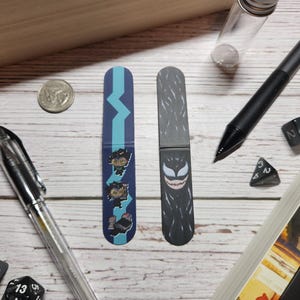 May include: Two colorful bookmarks. One bookmark features a blue and teal design with a graphic of three cartoon characters. The other bookmark is black with a graphic of a black and white creature with a large mouth.