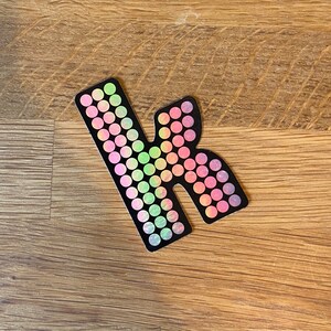 May include: A black letter "k" sticker with a gradient of pink, green, and yellow circles. The sticker is on a wooden surface.