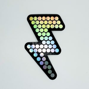 May include: Black lightning bolt sticker with a holographic, rainbow-colored dot pattern. The sticker is a stylized representation of a lightning bolt, with a solid black outline and a series of small, iridescent circles filling the interior.