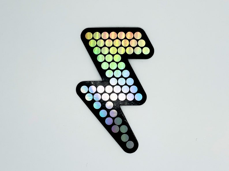 May include: Black lightning bolt sticker with a holographic, rainbow-colored dot pattern. The sticker is a stylized representation of a lightning bolt, with a solid black outline and a series of small, iridescent circles filling the interior.