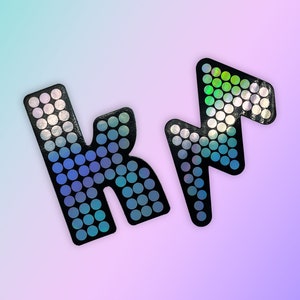 May include: Two black, glittery letter-shaped embellishments, a stylized "K" and a lightning bolt, are displayed against a gradient background of blue, purple, and pink. The letters are covered in small, iridescent circles.