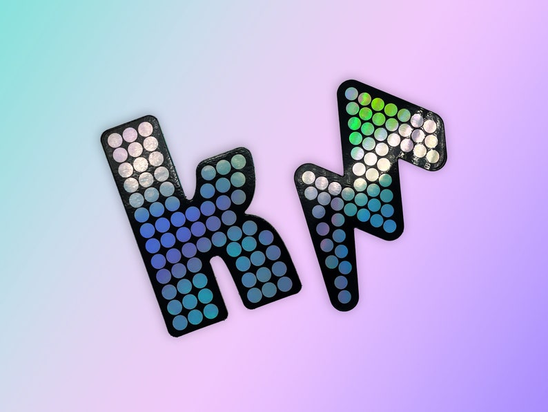May include: Two black, glittery letter-shaped embellishments, a stylized "K" and a lightning bolt, are displayed against a gradient background of blue, purple, and pink. The letters are covered in small, iridescent circles.