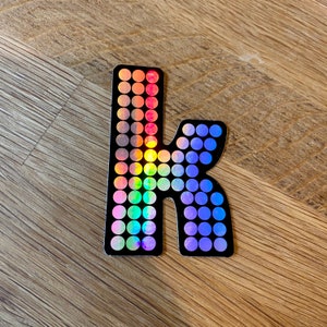 May include: A holographic sticker in the shape of the lowercase letter "k". The sticker is black with rows of colorful, iridescent circles. The colors transition from red to purple, creating a rainbow effect.