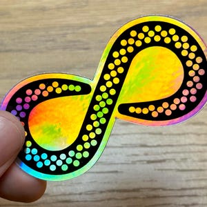 May include: A holographic sticker with a black infinity symbol outlined with a row of white dots. The background of the sticker is a rainbow of colors, including yellow, green, blue, and purple.