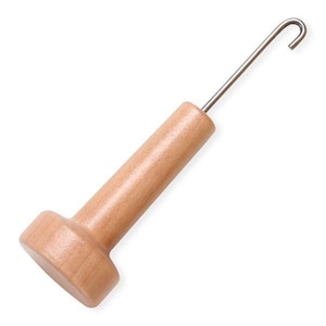 May include: A rug hooking tool featuring a smooth, light-toned wooden handle with a cylindrical grip and a wider, flat base. A thin, silver-colored metal shaft extends from the top of the handle, terminating in a small, curved hook at the end.