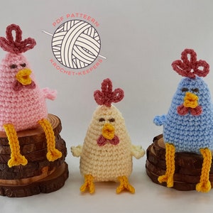 May include: Three crocheted chicken amigurumi toys. One is pink, one is blue, and one is cream. They are all sitting on wooden rounds.