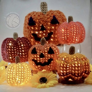 May include: Five crocheted pumpkin decorations with light inside. The pumpkins are orange, yellow, and pink. The pumpkins are all different sizes and have different facial expressions. The pumpkins are sitting on a table with a sunflower in front of them.