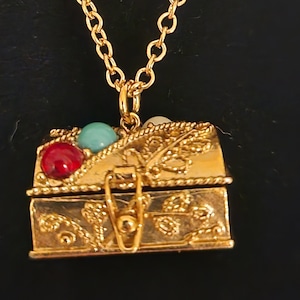 May include: A gold-tone necklace with a small, ornate treasure chest pendant. The chest is decorated with red, turquoise, and white stones and has a hinged lid.