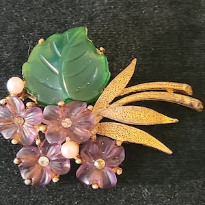 以下が含まれることがあります： 緑の葉と紫の花が付いたゴールドトーンのブローチ。花の中央にはラインストーンが飾られています。