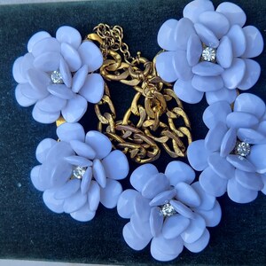 Pode incluir: Um colar com cinco pingentes de flores lilás claras e uma corrente dourada. Cada flor tem um strass central. O colar é exibido em uma superfície de veludo verde escuro.