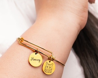 May include: A gold bangle bracelet with two charms. One charm is a round disc with the word "Honey" engraved on it. The other charm is a round disc with a cat's face engraved on it.