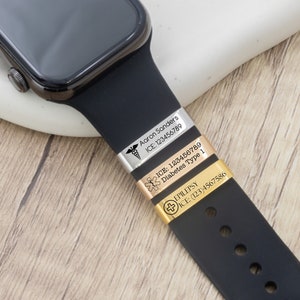 K&ouml;nnte beinhalten: Eine schwarze Smartwatch mit schwarzem Armband, versehen mit drei medizinischen Notfallarmb&auml;ndern. Die B&auml;nder sind silber, gold und ros&eacute;goldfarben und mit medizinischen Informationen graviert, einschlie&szlig;lich Namen, Erkrankungen und Notfallnummern. Die Uhr liegt auf einer wei&szlig;en Oberfl&auml;che.