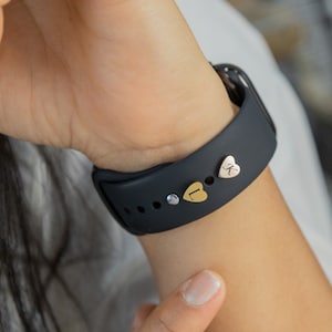 May include: A black Apple Watch band with three small heart-shaped charms. The charms are gold, silver, and rose gold and each has a letter engraved on it.