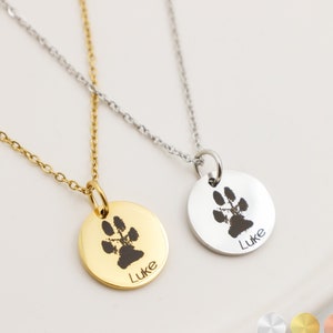 May include: Two gold and silver necklaces with round pendants featuring a paw print and the name "Luke" engraved on each. The necklaces are on a white background with three small circles showing the different metal options: silver, gold, and rose gold.