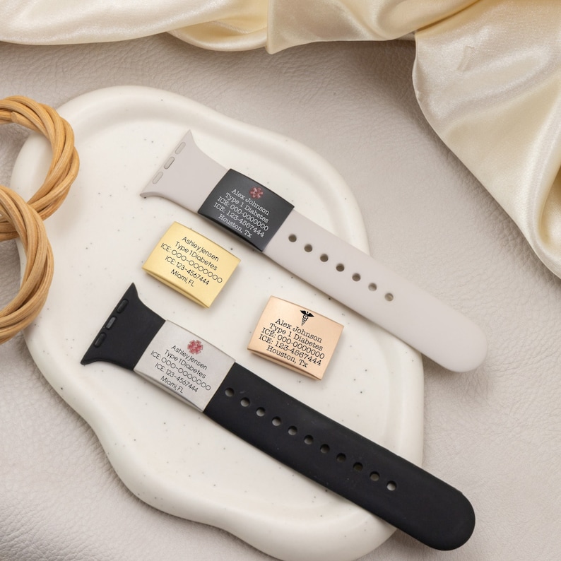 K&ouml;nnte beinhalten: Zwei schwarze und wei&szlig;e Apple Watch-Armb&auml;nder mit medizinischen Warnhinweisen. Die Anh&auml;nger sind mit Namen, medizinischen Bedingungen und Kontaktinformationen graviert. Die Armb&auml;nder liegen auf einer wei&szlig;en Oberfl&auml;che.