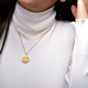 May include: A gold chain necklace with a round pendant featuring a dog's face. The necklace is worn by a person wearing a white turtleneck sweater.
